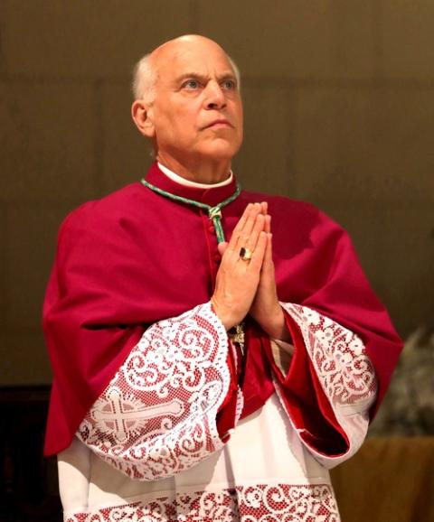 The archbishop wears choir dress, hands folded and head raised in prayer. 
